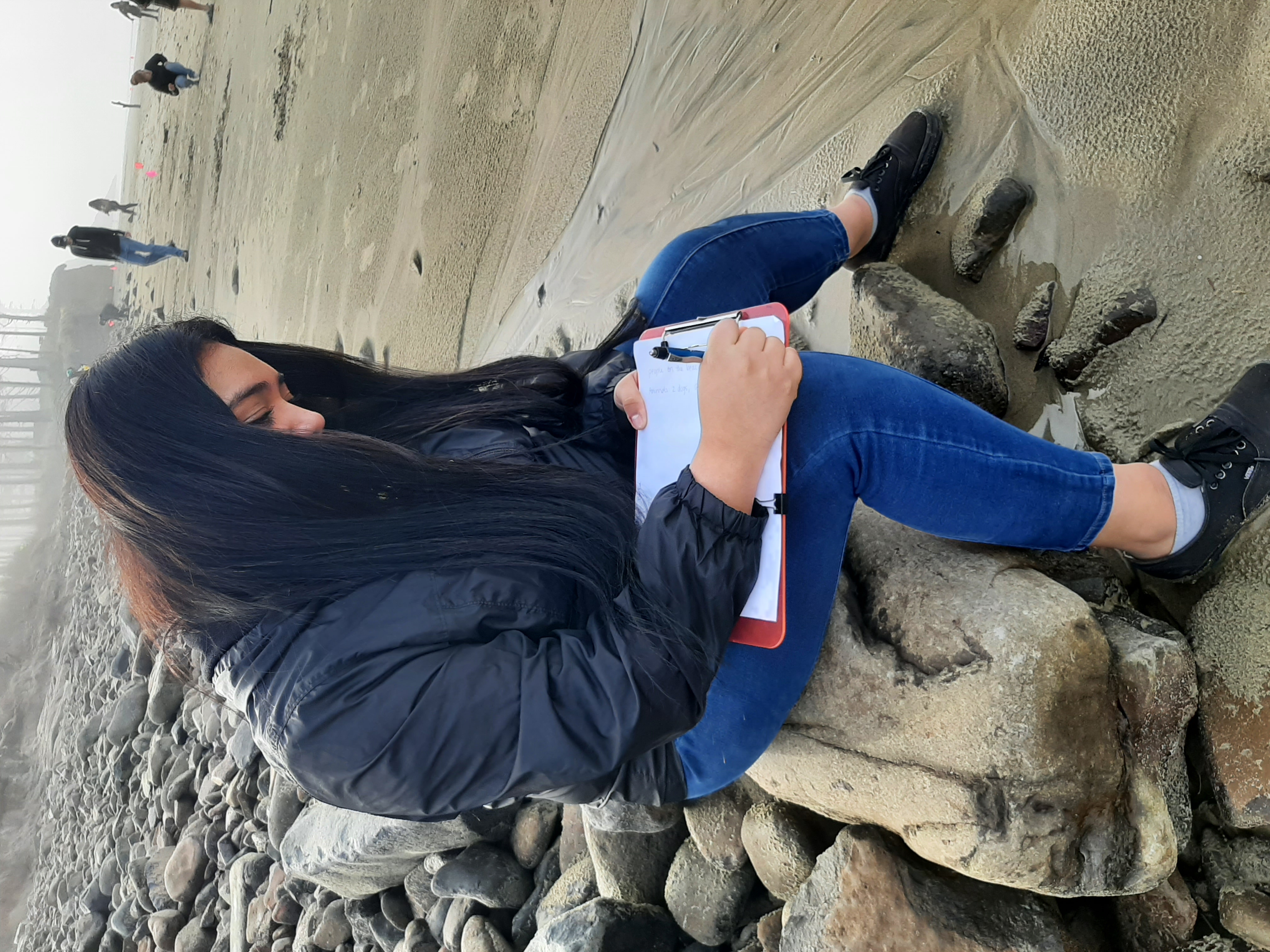 A student with long, black hair sits on a rock and records data. 