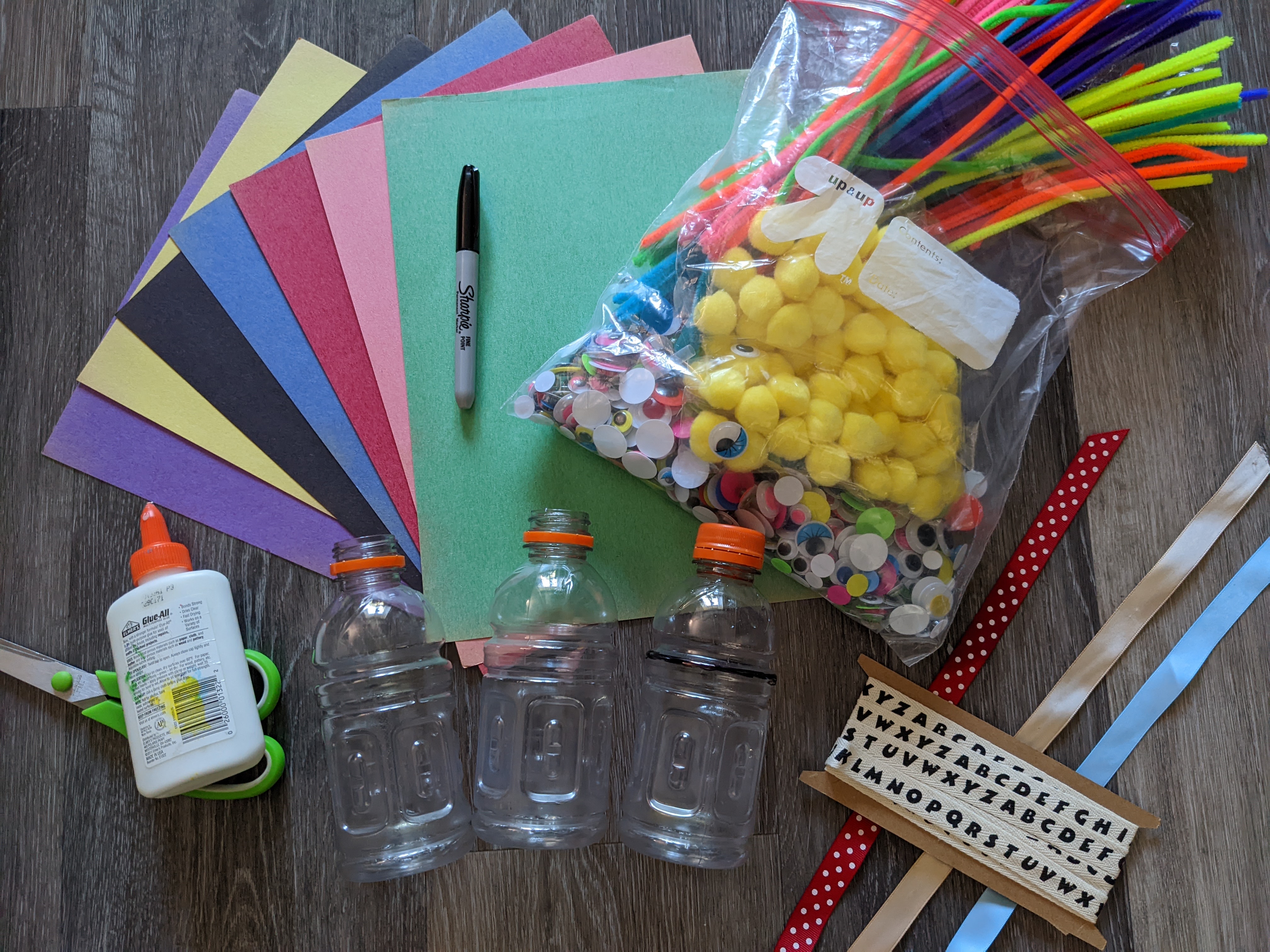 Various craft supplies including colored construction paper, plastic bottles, ribbon, pipecleaners, and googly eyes spread out on a table.