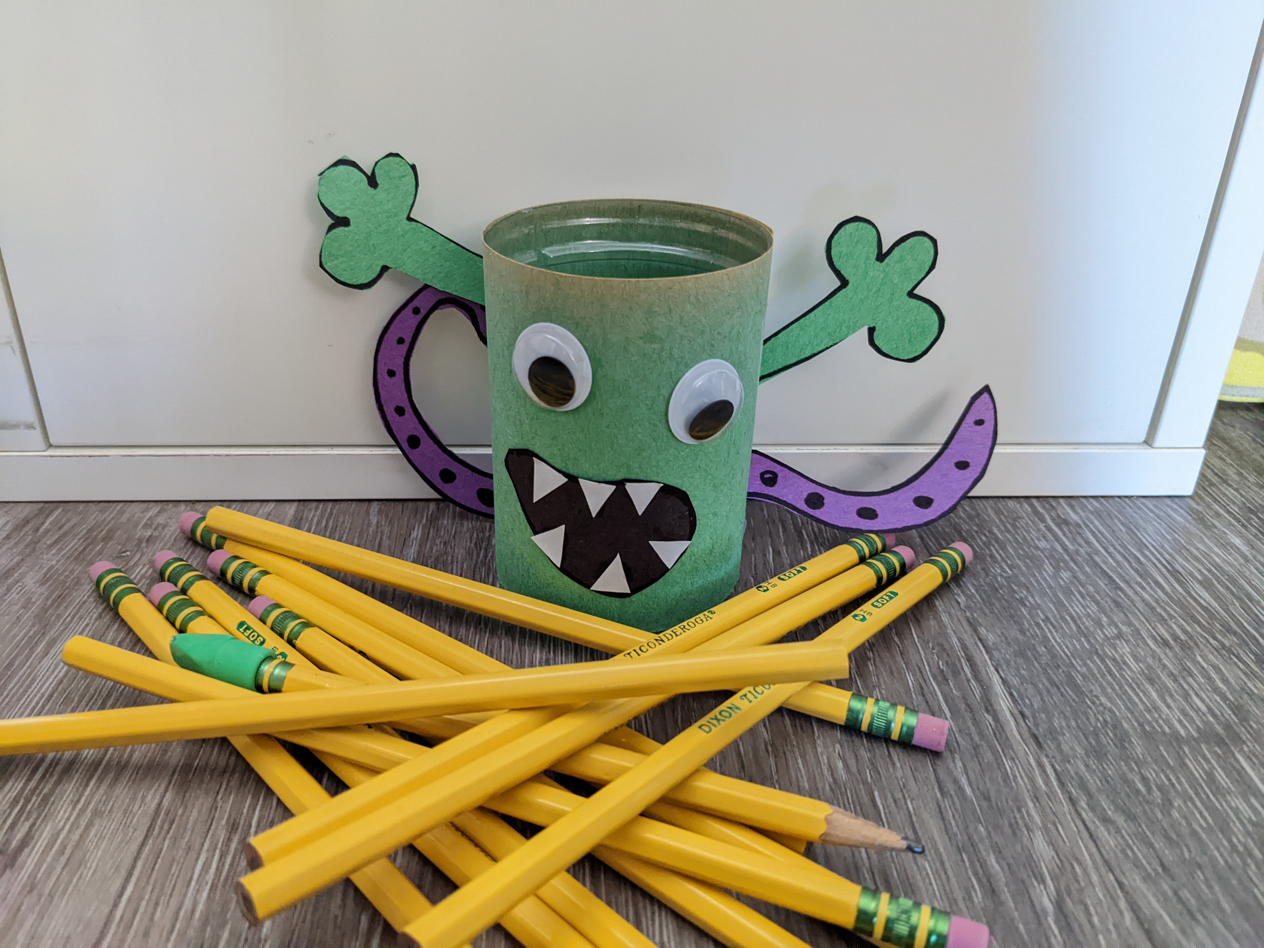 A green pencil cup decorated like a monster in front of a pile of pencils.