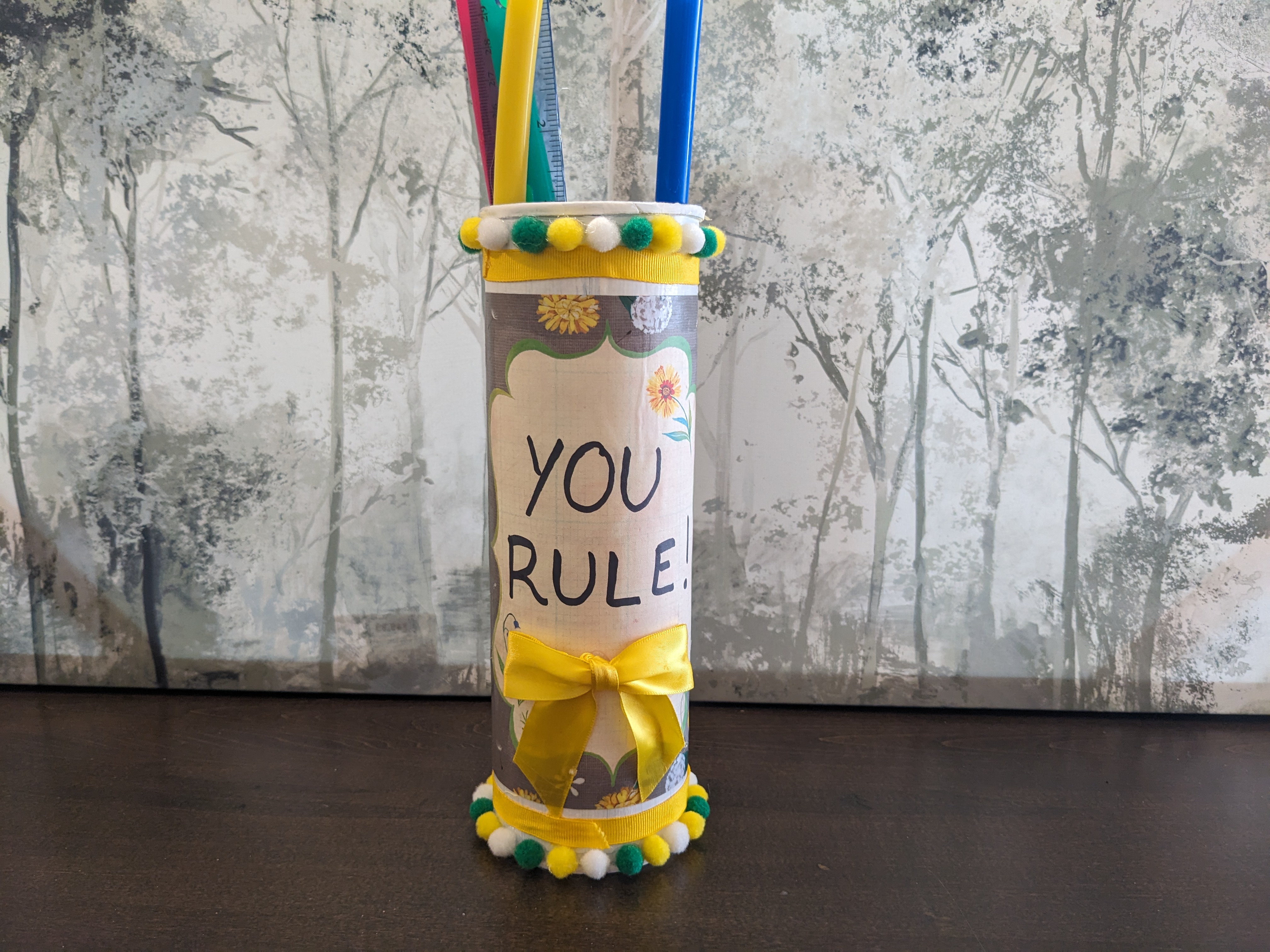 A tall canister decorated and holding a ruler and long straws.