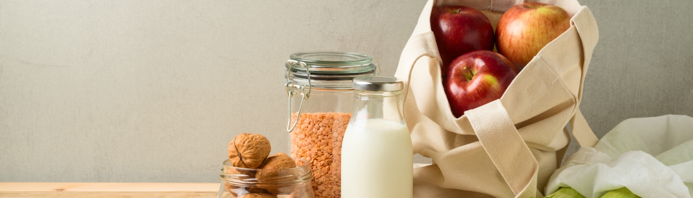 A cloth bag filled with apples and zucchini, a glass jar of milk, a glass container of lentils, and a glass jar of walnuts sitting on a wooden table.