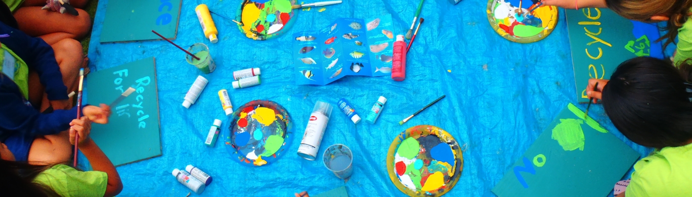 Students participating in an educational marine debris art activity. The students are in a circle around a plastic tarp, painting marine debris messages on pieces of wood.