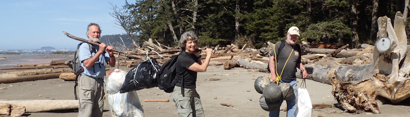 Three people walk on a beach on the Olympic Coast carrying plastic bags full of garbage.