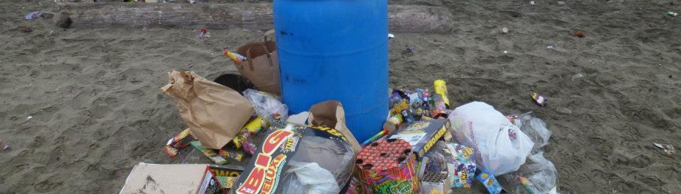 A trash can overflowing with trash on a beach with more litter around it.