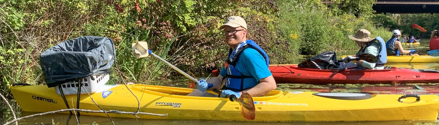 A person paddling a kayak smiles for a picture. A laundry basket lined with a black garbage bag has been attached to the front of the kayak.