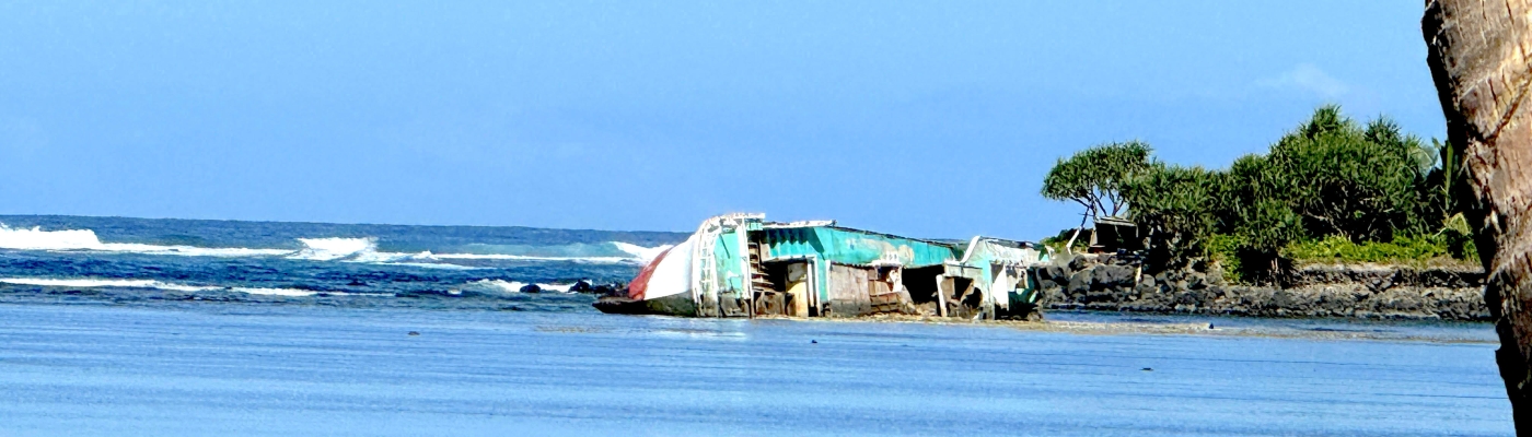 A dilapidated vessel on its side in shallow water near a rocky shoreline.