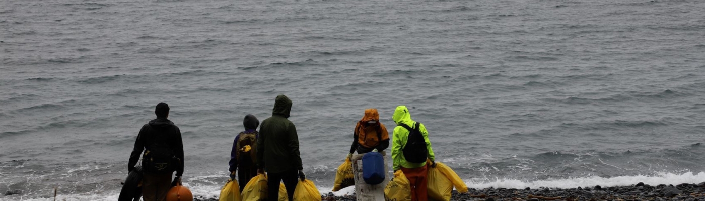 A crew of people holding filled garbage bags standing at the shoreline and watching an incoming small boat.