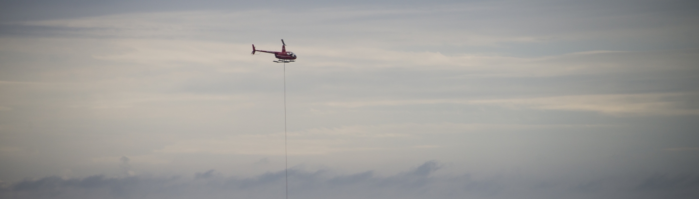A helicopter carrying debris hovers over a flat barge sitting on the ocean that is piled with bags of debris.