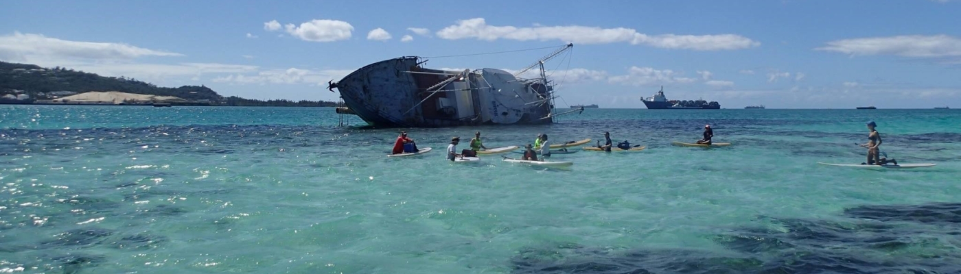 A derelict vessel in the background while several people swim in a beautiful lagoon.