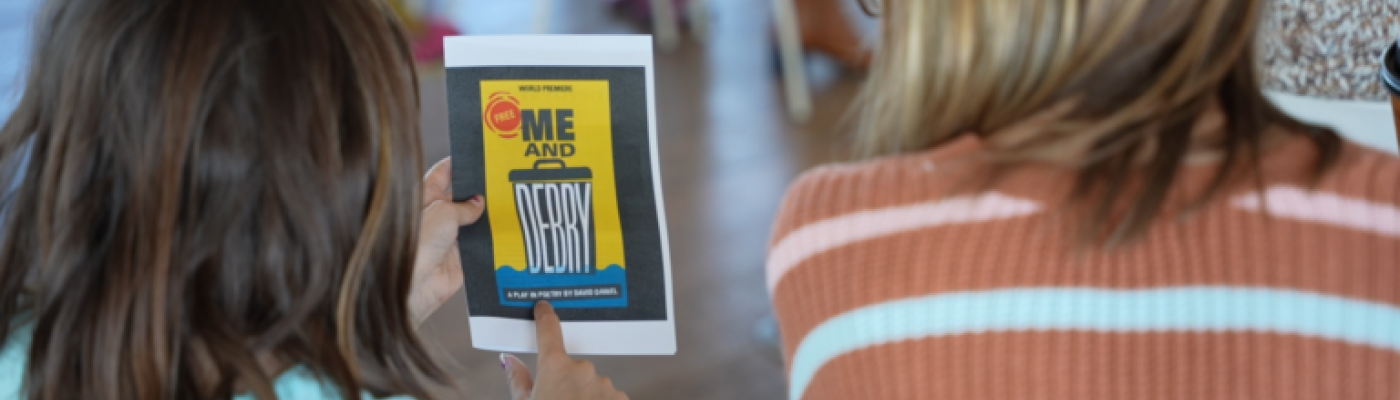 A woman with blond hair and a child with brown hair, seen from behind as they are reading the cover of the playbill for “Me and Debry.” 