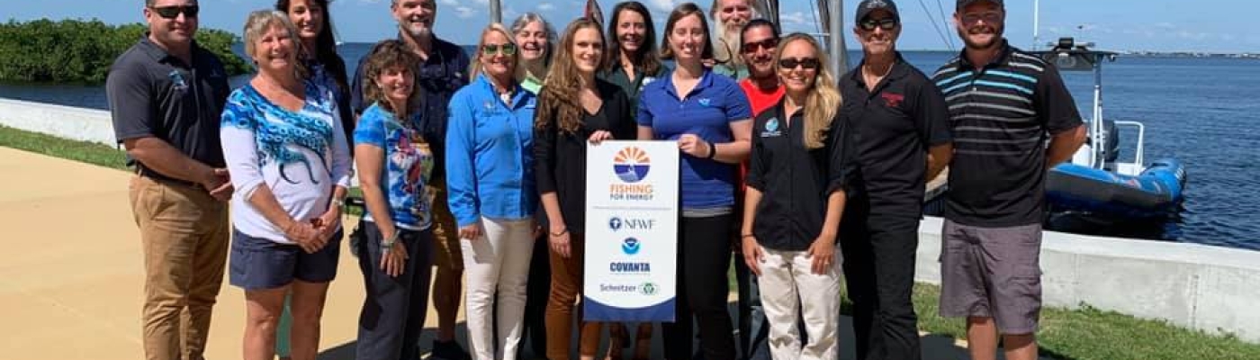 15 people stand together for a picture holding up a small sign that says "Fishing for Energy".