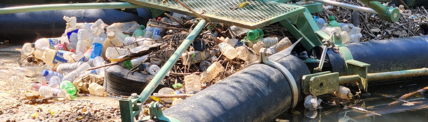 Plastic bottles and other plastic trash items mixed with leaf litter collect behind the black piping of an interception device in a shallow, muddy waterway.