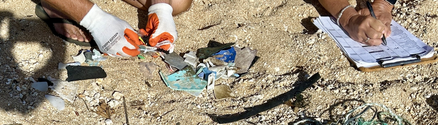 Two people's hands are seen examining and documenting small plastic pieces and netting on a data card on a sandy beach.