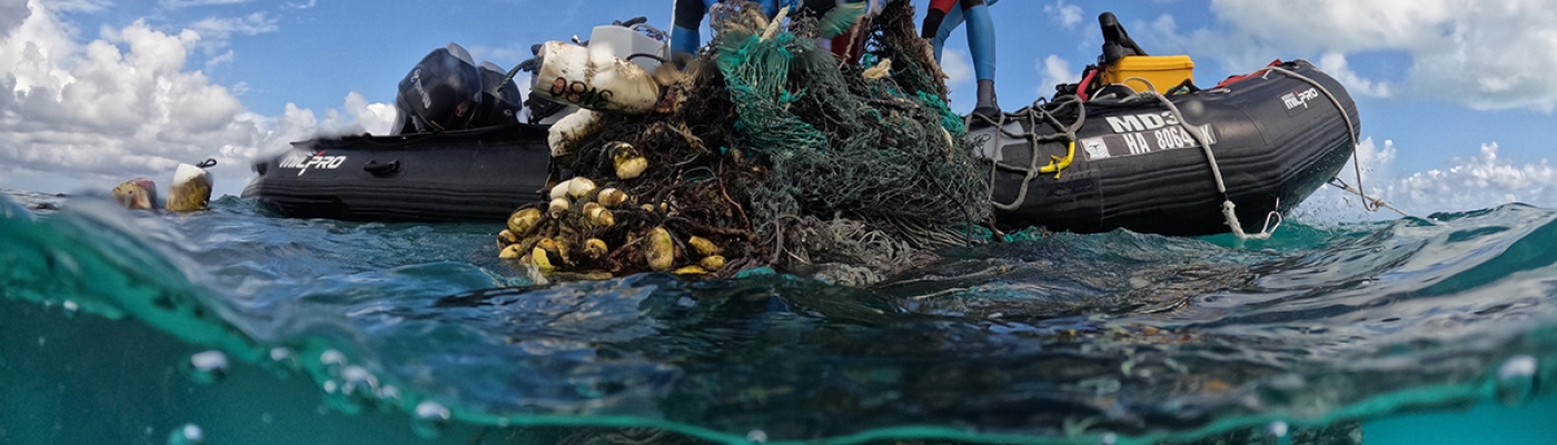 Three people stand on a small boat and are seen pulling a large mass of nets out of the water onto the vessel. 