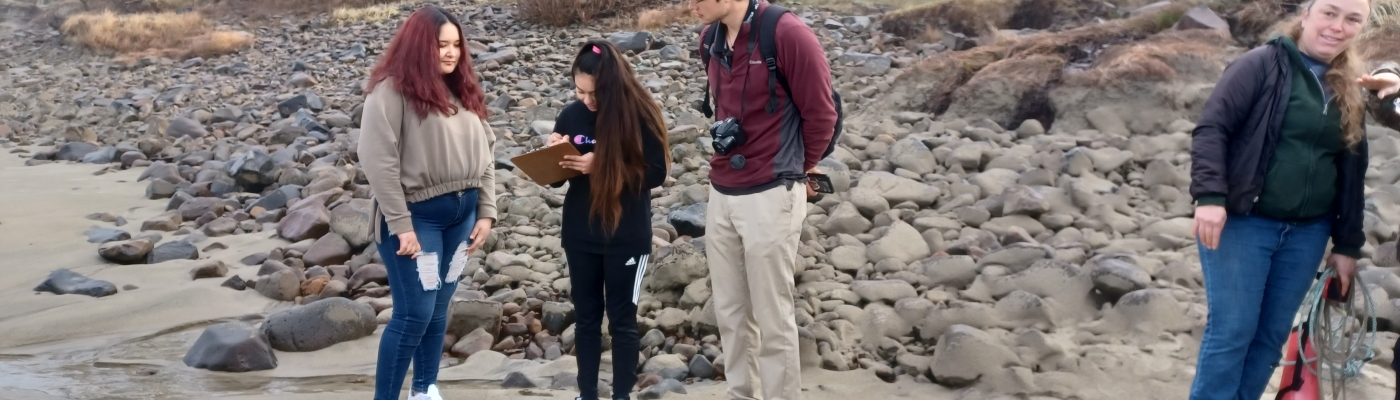 Two students and their teacher review a clipboard on a rocky beach.