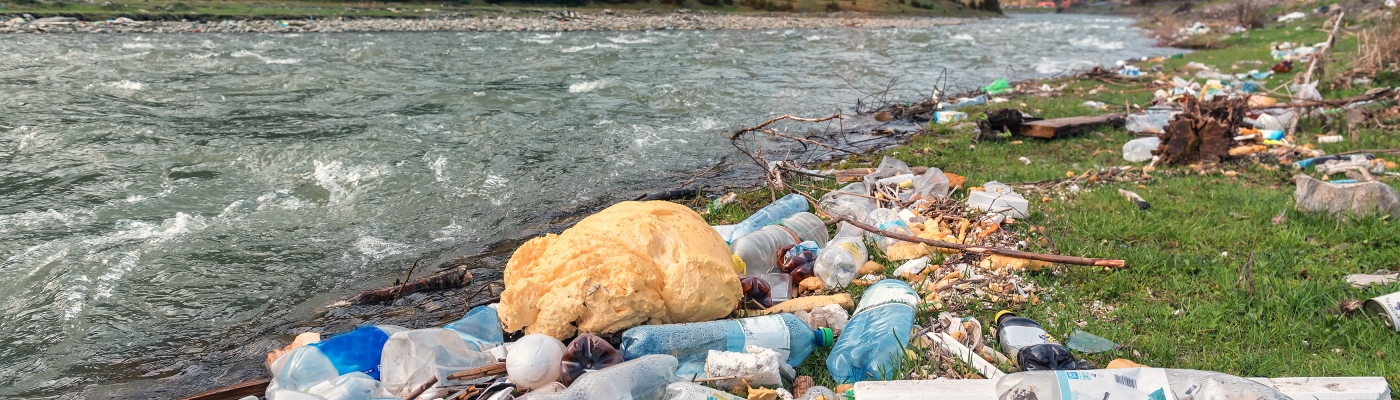 Plastic marine debris items collected on the shoreline of a river as the sun sets behind mountains.