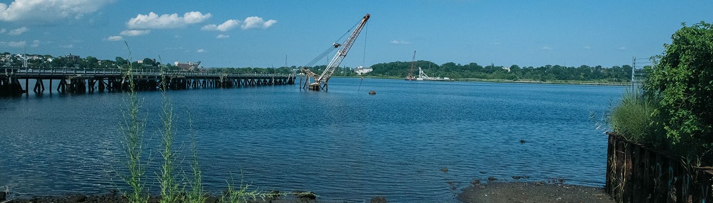 A crane sticks out of the water next to a pier on a sunny day as seen from a rocky, muddy urban shore.