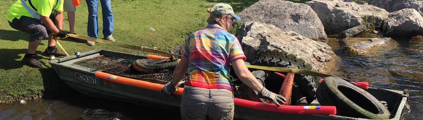 People pulling a boat ashore that is filled with marine debris collected along a lake shoreline.