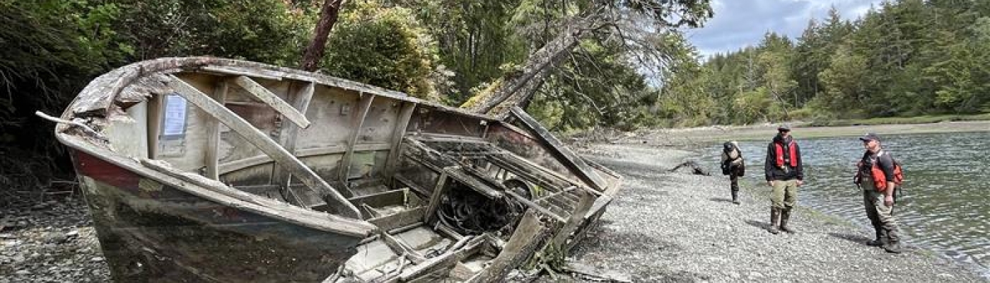 A broken wooden boat lays on it side on a rocky, tree-lined shoreline with three government officials in the background. 