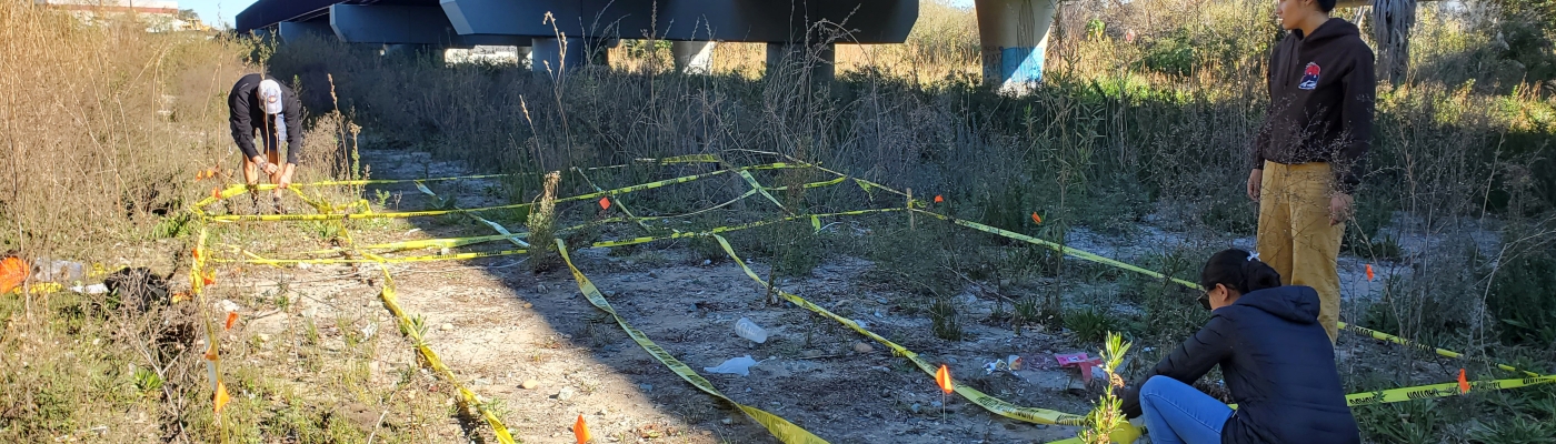 Three researchers use yellow tape to set up a grid covering bare and grassy vegetation for observing marine debris via uncrewed aerial systems near a railroad bridge overpass.