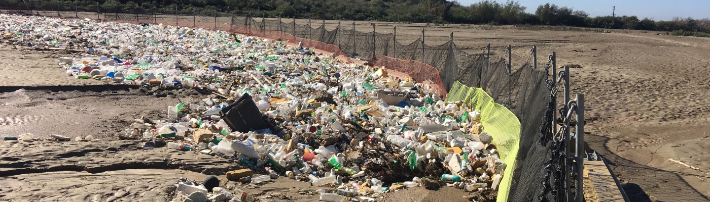 A large trash boom goes from one side of a dry river bed to the other with lots of trash stuck on one side.