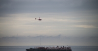 A helicopter carrying debris hovers over a flat barge sitting on the ocean that is piled with bags of debris.
