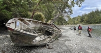A broken wooden boat lays on it side on a rocky, tree-lined shoreline with three government officials in the background. 