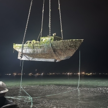 A derelict vessel being hoisted out of the water with a series of chains at night with a city skyline illuminated in the distance.