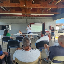 An outdoor classroom setting with a group of people sitting in chairs listening to a presenter standing in front of a projector.