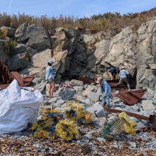 People filling large, white bags with marine debris on a rocky island coastline on a sunny day.