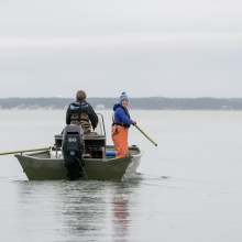 Two people stand on a boat holding long poles that will be used to pull a crab trap up out of the water.