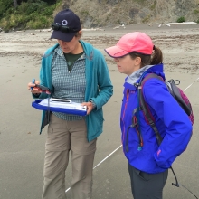 Two people looking at a clipboard and GPS tracker on a beach.