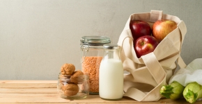 A cloth bag filled with apples and zucchini, a glass jar of milk, a glass container of lentils, and a glass jar of walnuts sitting on a wooden table.