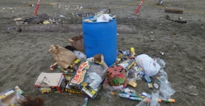 A trash can overflowing with trash on a beach with more litter around it.