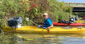 A person paddling a kayak smiles for a picture. A laundry basket lined with a black garbage bag has been attached to the front of the kayak.