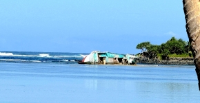 A dilapidated vessel on its side in shallow water near a rocky shoreline.