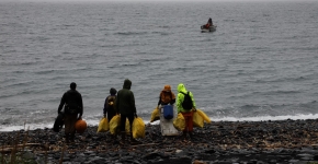 A crew of people holding filled garbage bags standing at the shoreline and watching an incoming small boat.