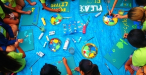 A group of students painting "Please Recycle" messages on a blue sign.