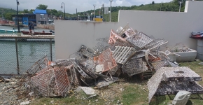 A pile of broken fishing traps sit in the grass in front of a wall and fence with harbor docks in the background.