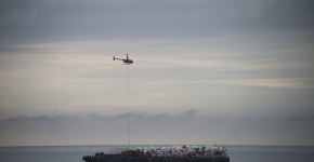 A helicopter carrying debris hovers over a flat barge sitting on the ocean that is piled with bags of debris.