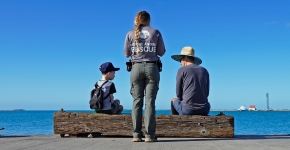 A Hawaii Marine Animal Response staff member speaking to a child and adult sitting on timber overlooking the ocean. 