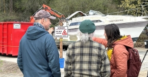 Three people huddle together and talk while facing a boat getting demolished in a gravel parking lot.