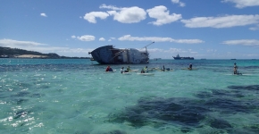 A derelict vessel in the background while several people swim in a beautiful lagoon.