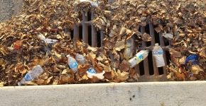 Plastic water bottles and chip bags mixed with leaf litter sit on top of the grate of a storm drain. 