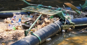 Plastic bottles and other plastic trash items mixed with leaf litter collect behind the black piping of an interception device in a shallow, muddy waterway.