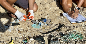 Two people's hands are seen examining and documenting small plastic pieces and netting on a data card on a sandy beach.