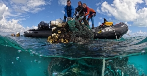 Three people stand on a small boat and are seen pulling a large mass of nets out of the water onto the vessel. 