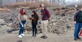 Two students and their teacher review a clipboard on a rocky beach.