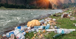 Plastic marine debris items collected on the shoreline of a river as the sun sets behind mountains.