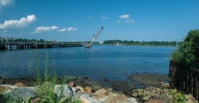 A crane sticks out of the water next to a pier on a sunny day as seen from a rocky, muddy urban shore.